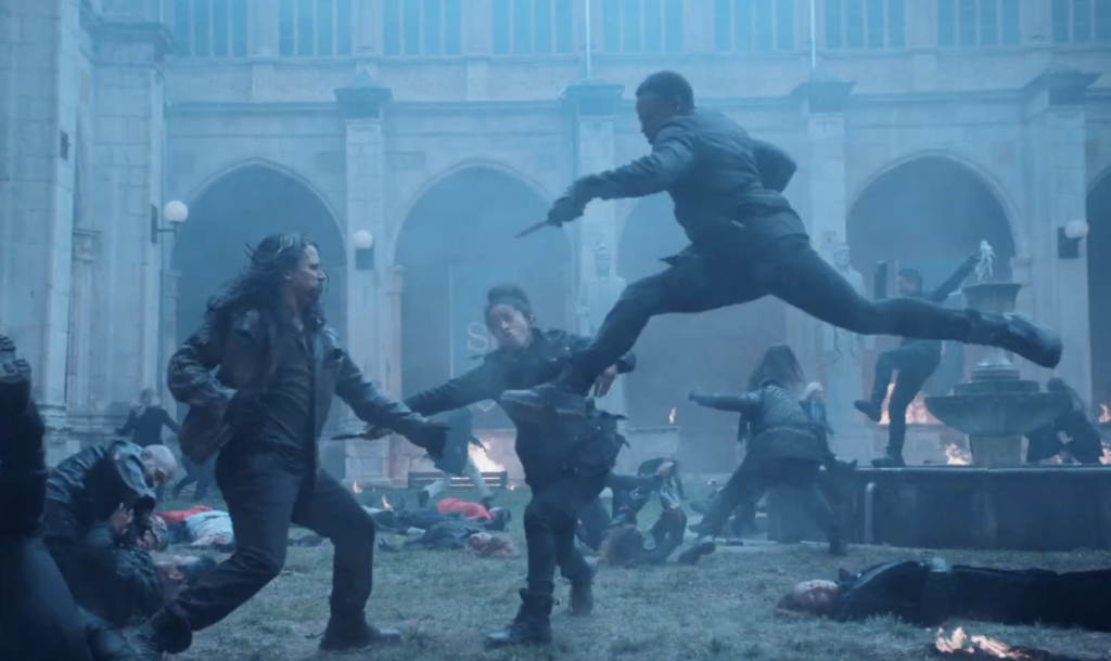 Scene at St. Jude with blue, hazy background. Janine Hathaway leaping through the air during the strigoi attack with her stake pointed at one and another Guardian behind the same strigoi striking forward